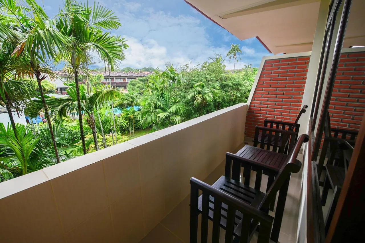 A private balcony with dark wooden chairs looks out over palm trees and tropical greenery toward low hotel buildings.