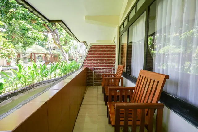 A covered balcony with wooden chairs beside glass doors and sheer curtains, overlooking a garden with trees and tropical plants.