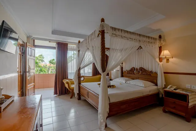 A bedroom with a wooden canopy bed draped in white netting, a yellow bench, a wall-mounted television, and an open door onto a balcony with palm trees outside.
