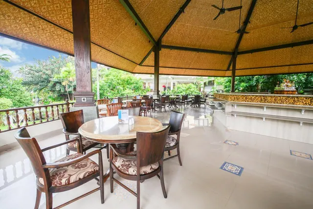 A covered open-sided dining pavilion with round wooden tables, upholstered chairs, ceiling fans, and a bar counter on the right, with garden trees visible outside.