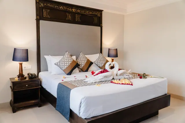 A made-up king bed with white linen, towel swans and flower petals, flanked by two bedside lamps and a carved dark wood headboard.