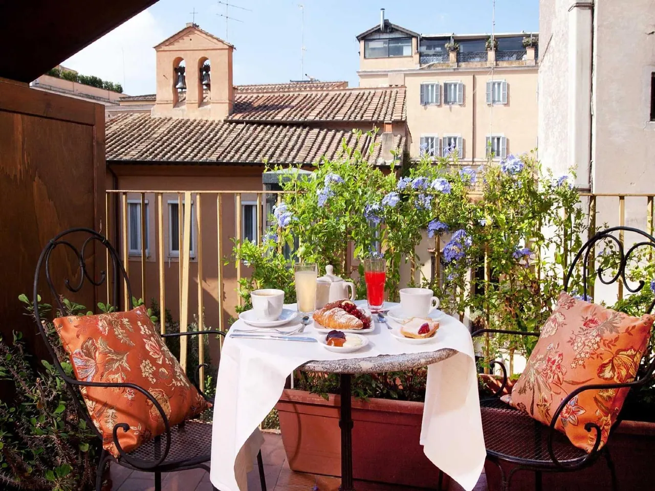 A breakfast table with coffee cups, pastries, juice and a jam dish sits on a balcony overlooking rooftops and a bell tower.