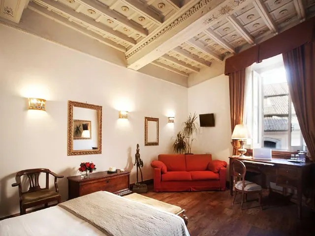 A bedroom with a coffered ceiling, a bed, a red sofa, a wall mirror and a desk by a window.