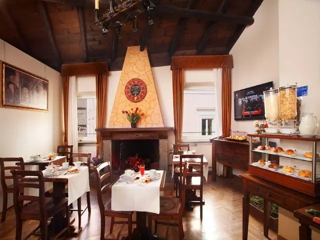 A breakfast room with wooden tables set with white cloths and cups, a fireplace, a beamed wooden ceiling, a wall-mounted television and a pastry display on the right.