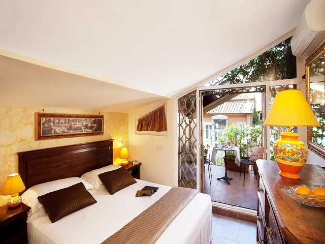 A bedroom with a double bed, bedside lamps, a wooden dresser and an open glass door onto a terrace with a table and chairs.
