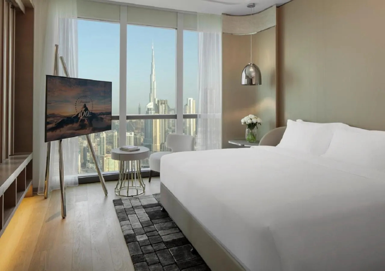 A bedroom with a large white bed, a floor-standing television, and floor-to-ceiling windows looking out at the Burj Khalifa and surrounding towers.