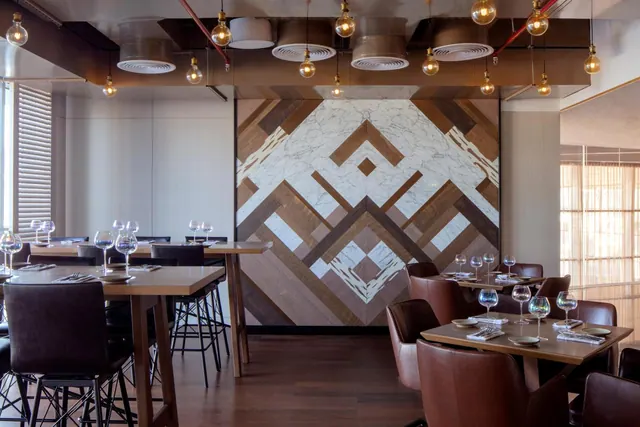 A restaurant dining room with wooden tables, leather chairs, pendant bulbs and a wall of wood and marble inlay.