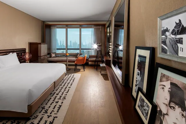 A bedroom with a large white bed, a seating area by floor-to-ceiling windows, and a city skyline visible outside.