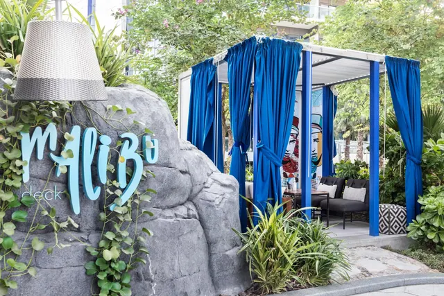A blue-curtained cabana with grey sofas and cushions stands beside a stone wall bearing the Malibu deck sign, surrounded by garden plants.