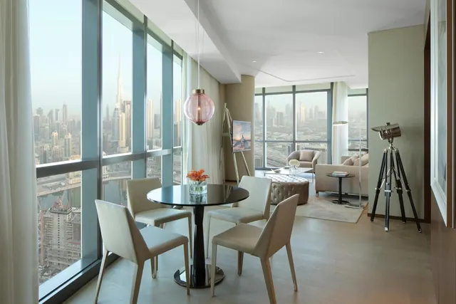 A living and dining area with floor-to-ceiling windows overlooking a city skyline that includes the Burj Khalifa.