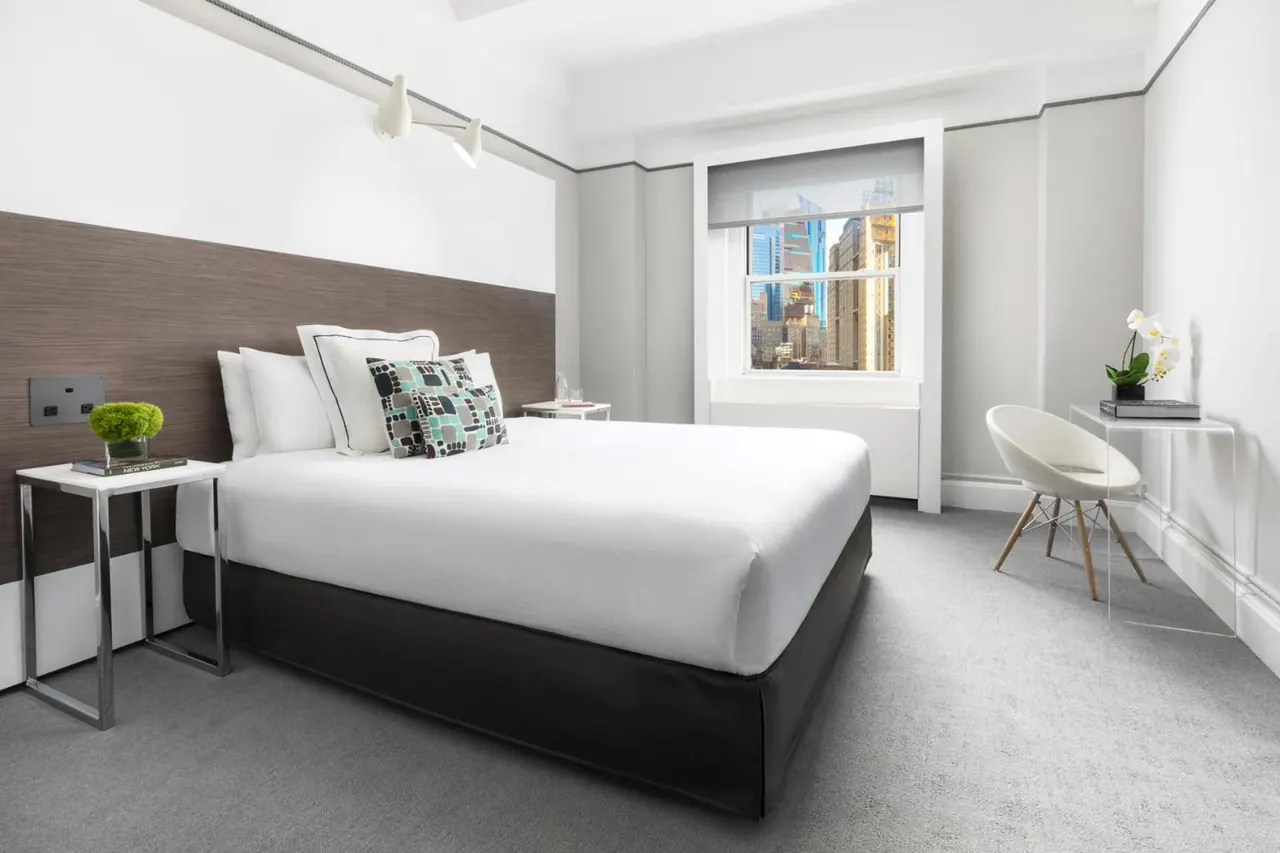 A white bedroom with a made bed, a window showing city buildings, and an acrylic desk with a chair beside it.