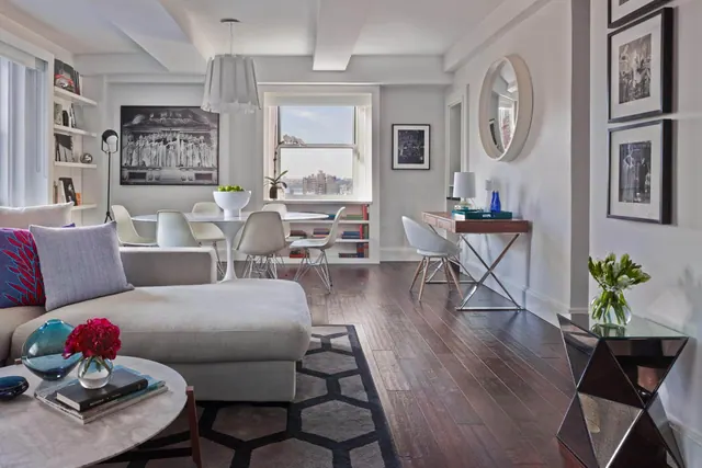 A bright living area with a grey sofa, a round white dining table with chairs, built-in bookshelves, and a window showing nearby city buildings.