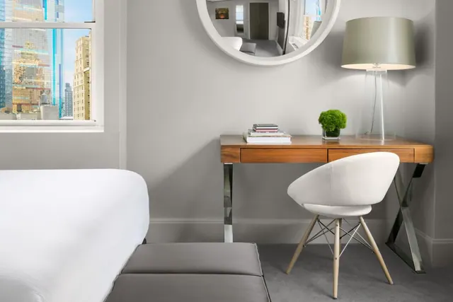A white bed, a grey bench, and a wooden desk with a white chair and table lamp sit in a bedroom beside a window overlooking tall city buildings.