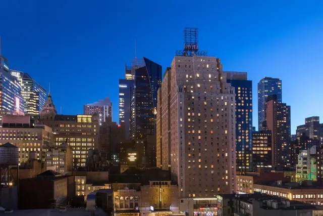 Twilight view of a city skyline with a tall tan hotel tower topped by a rooftop HOTEL sign and illuminated office towers behind it.