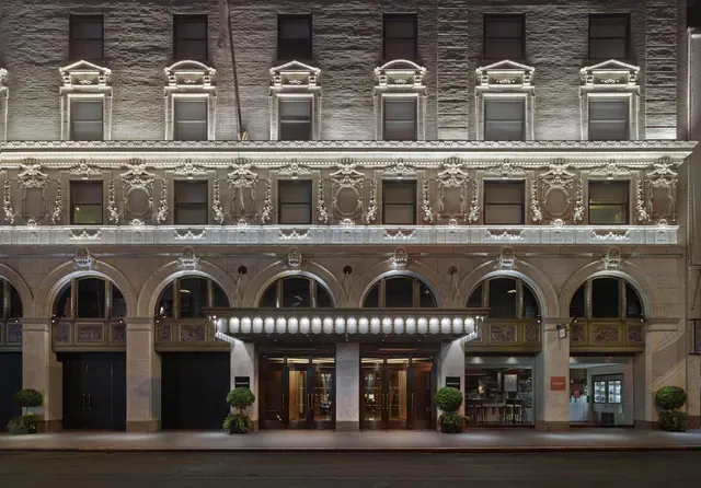 Night view of an ornate stone building facade with an illuminated entrance canopy and trimmed topiary plants beside the doors.