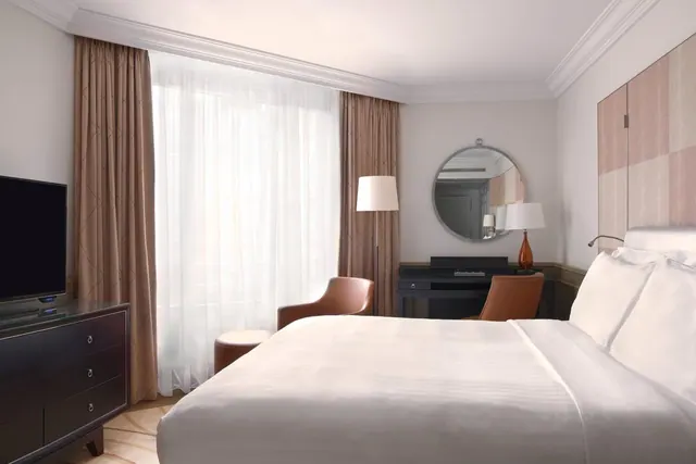A bright hotel bedroom contains a large bed, seating, a desk, television and tall curtained windows.