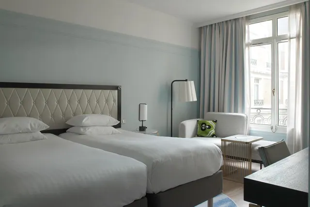 A bright bedroom with twin beds, a seating area, a desk and tall windows overlooking the street.