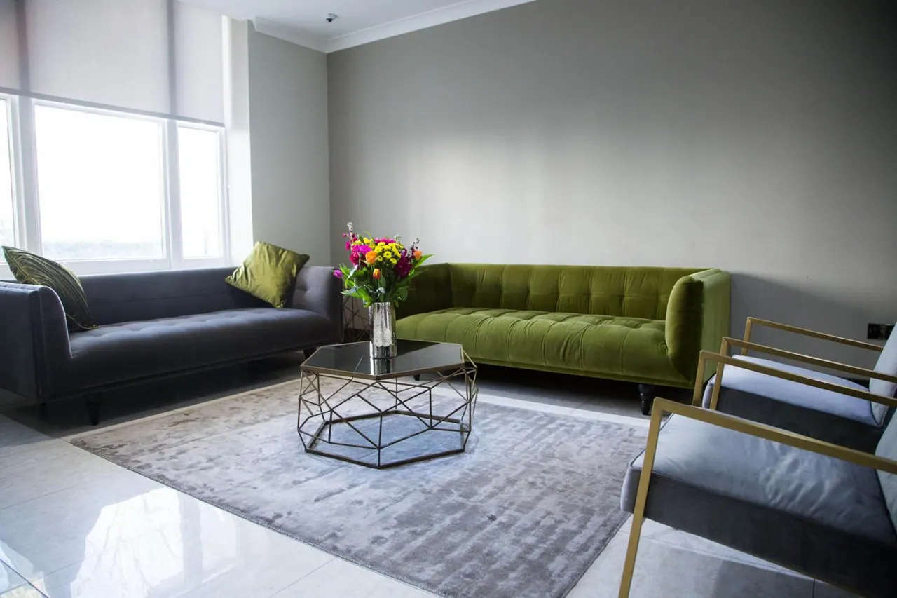 A seating area with a grey sofa, a green velvet sofa, a geometric coffee table with a vase of flowers, and gold-framed armchairs on a grey rug.