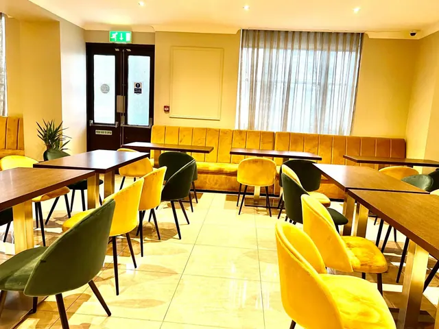 A dining room with a yellow velvet banquette along the back wall, green and yellow upholstered chairs and wooden tables on a cream tiled floor.