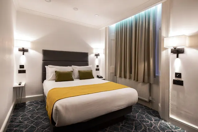 A bedroom with a double bed under a yellow throw, dark upholstered headboard, green cushions, wall-mounted lights and closed curtains at the window.