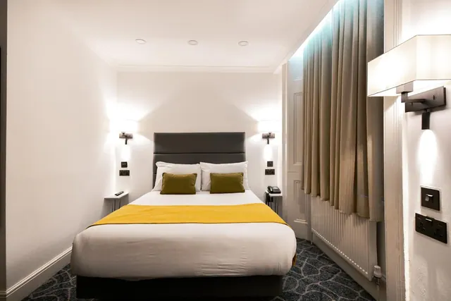A hotel bedroom with a double bed dressed in white linen, a yellow throw, olive cushions, wall-mounted reading lights on both sides, and beige curtains along the side wall.