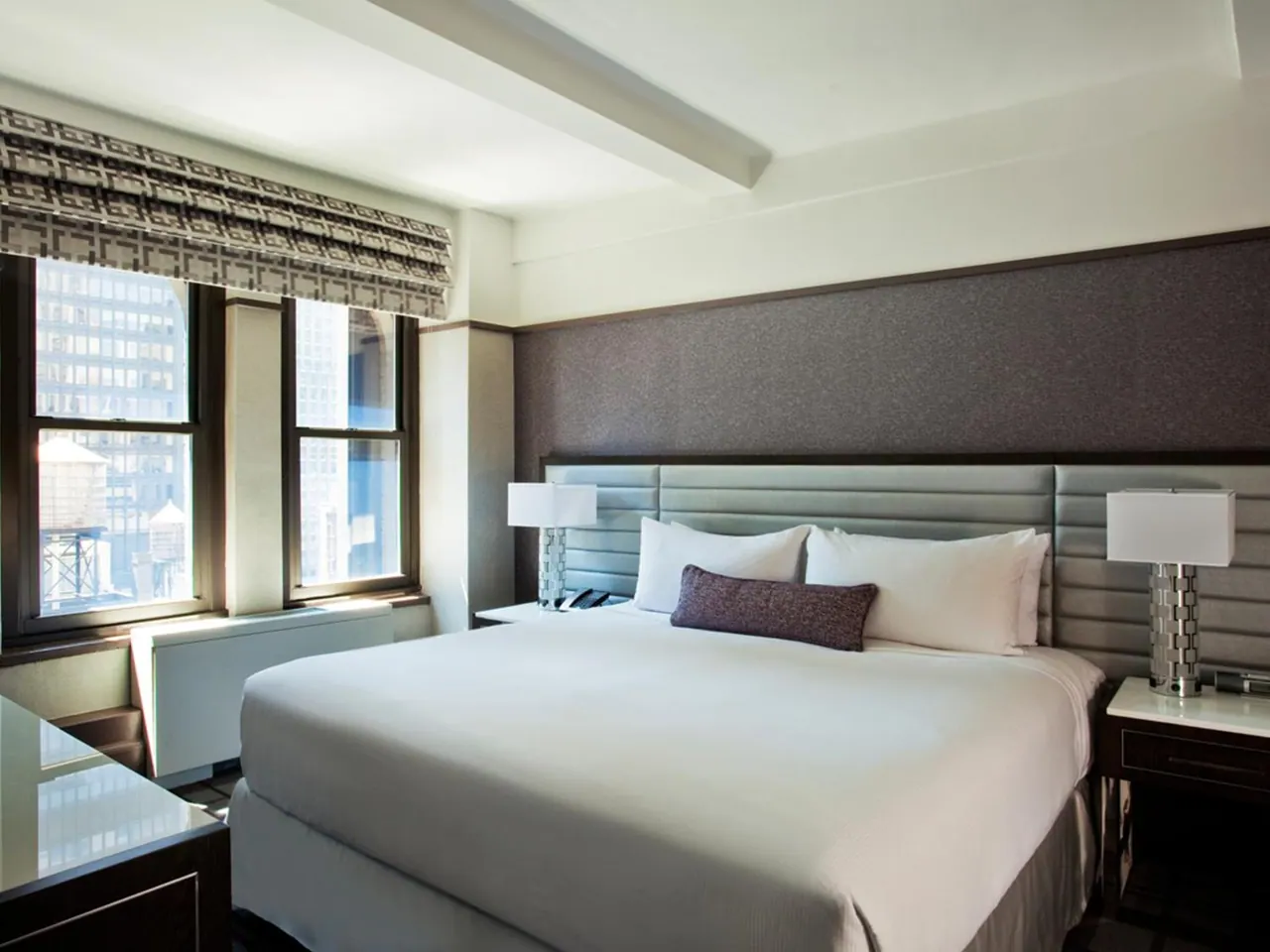 A made king bed with white linens and a purple pillow sits between two bedside lamps, with windows beside it looking out onto city buildings.