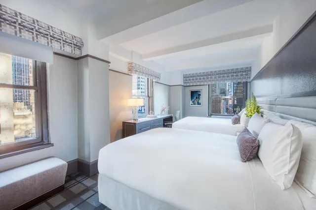 A hotel bedroom with two double beds, a desk with a lamp, and windows overlooking city buildings.