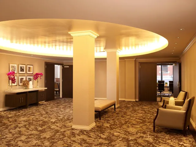 A lobby seating area with an illuminated circular ceiling, tall columns, upholstered armchairs and a bench, and a console table with pink orchids and framed prints.