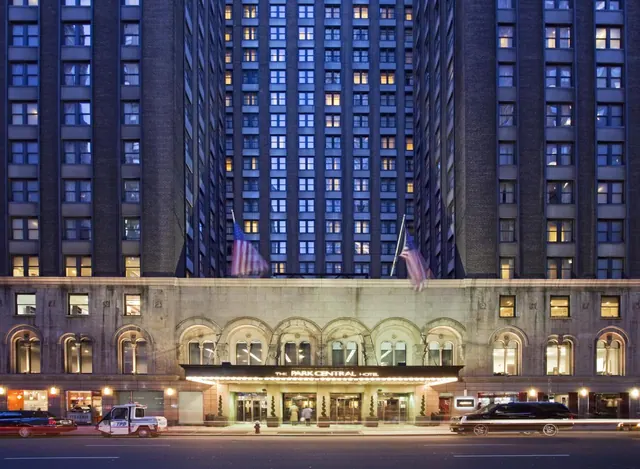 Dusk exterior of The Park Central Hotel, showing its tall windowed tower, stone-arched entrance under an illuminated canopy sign, two flags, and vehicles on the street.