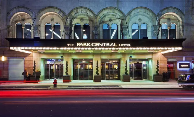 Exterior of The Park Central Hotel at night, with an illuminated entrance canopy, arched upper windows, and blurred red traffic light trails on the street.