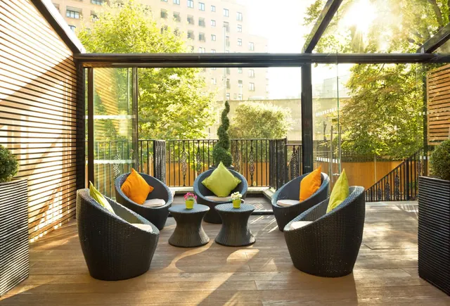 A covered terrace with wicker armchairs, cushions, two small side tables with potted flowers and topiary planters, with trees and a building visible beyond the glass walls.