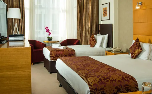 A hotel bedroom with two beds dressed in patterned red runners, two red armchairs, a wooden unit holding a television, and a pink orchid on a small round table by the window.