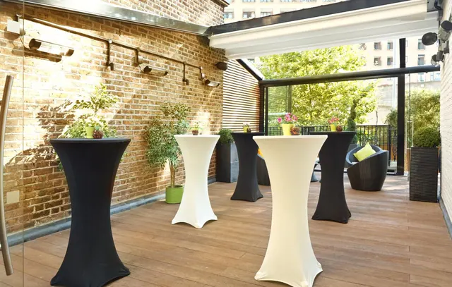 A covered outdoor terrace with a brick wall, wooden decking, high tables with black and white covers, wicker seating and potted plants, with trees and buildings visible beyond the glass.