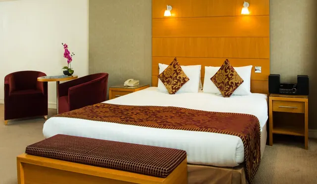 A double bed with a patterned runner and cushions, two armchairs beside a small round table with an orchid, and wooden bedside tables in a hotel room.