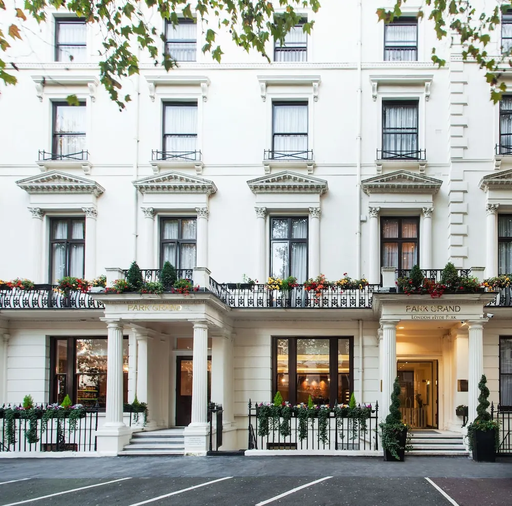The white stucco facade of the Park Grand hotel, with iron balconies, flower boxes, columned entrances and a tree branch in the upper frame, above an empty street with parking bays.