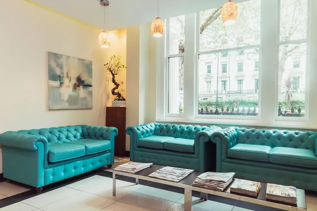 A lounge area with three teal tufted leather sofas, a glass coffee table holding newspapers, and tall windows looking out onto a street lined with trees and terraced buildings.