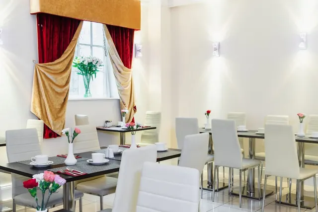 A bright dining room with white chairs and dark tables set with cups and saucers, flowers in vases, and red and gold curtains by a window.