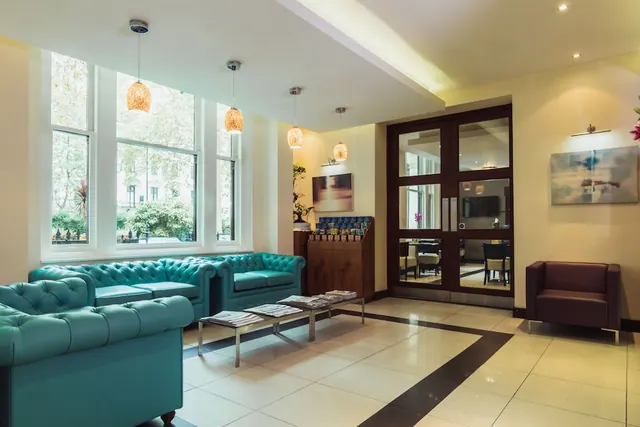 A lobby seating area with teal chesterfield sofas, low tables holding magazines, tall windows looking onto trees, and a dining room visible through glass doors.