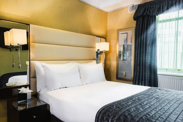 A bedroom with a made-up double bed with white pillows, a tall upholstered headboard, wall-mounted reading lamps, and dark curtains beside a window.