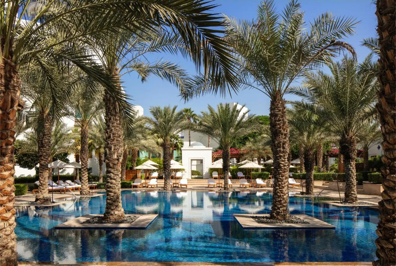 An outdoor pool lined with palm trees, white sun loungers and parasols, with a white building in the background.