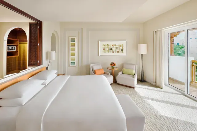 A bedroom with a large white bed, two armchairs, a side table and floor lamp, with sliding glass doors opening onto a balcony.