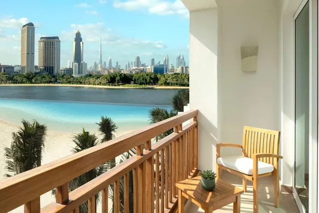 A private balcony with teak railings, a wooden chair with a white cushion and a small side table overlooks a sandy beach, water and the Dubai skyline.