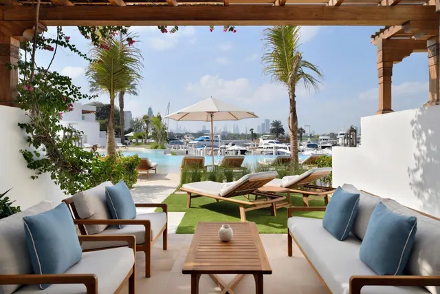 A covered terrace with cushioned sofas and a wooden coffee table overlooks a pool, sun loungers and a marina with yachts.