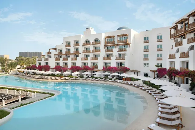 A large pool with white loungers and umbrellas sits in front of a white multi-storey hotel building with balconies and pink bougainvillea.