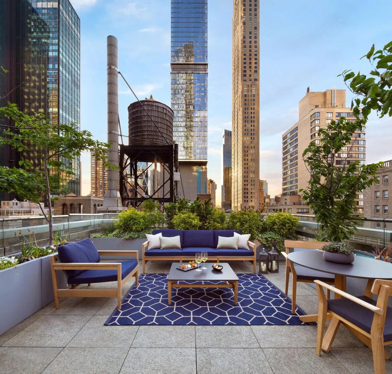 A rooftop terrace with navy outdoor sofa and armchairs, a patterned rug, a round dining table, and a view of city towers and a water tower.
