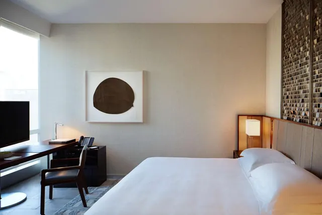 A bedroom with a made king bed, a framed abstract artwork on the wall, a desk with a television and chair, and a lit bedside lamp.