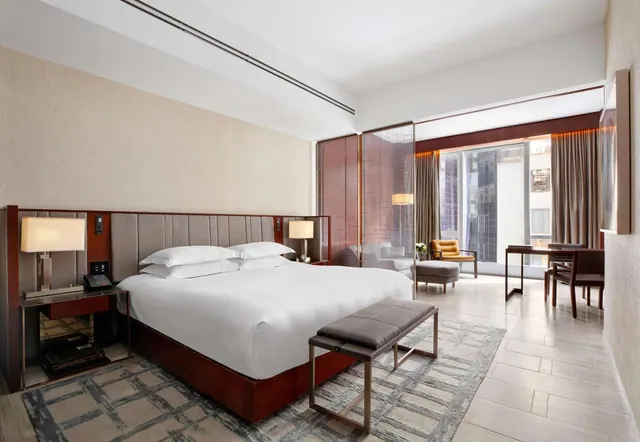 A hotel bedroom with a king bed, a bench at its foot, an armchair and ottoman by a large window showing city buildings, and a desk with chairs at right.
