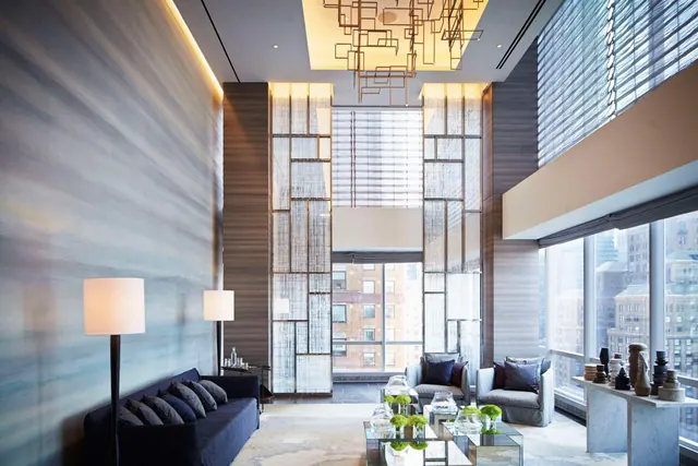A double-height living room with a dark sofa, two armchairs, floor lamps, glass coffee tables and large windows looking onto city buildings.