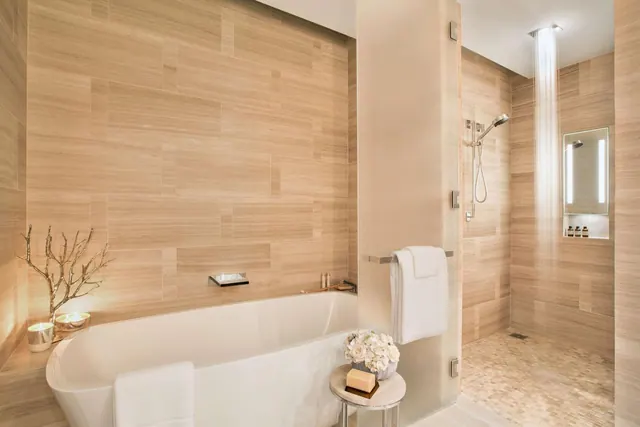 A bathroom with a freestanding white bathtub, a walk-in shower with an overhead rain shower, and beige stone-look wall tiles.
