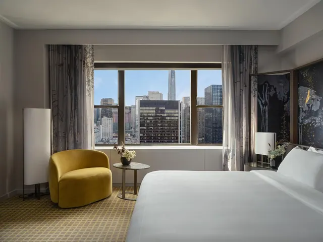 A bedroom with a made king bed, a yellow armchair and a large window looking out over a city skyline of tall buildings.